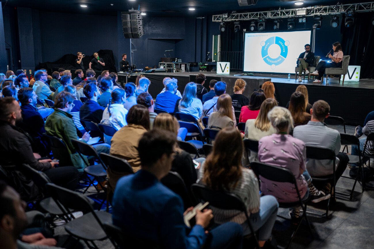 A large audience sits attentively in a dimly lit auditorium as two speakers on stage engage in a discussion. A presentation slide with a circular diagram is projected on a screen behind them