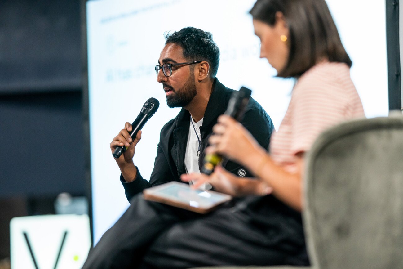 A man and woman are sitting and holding microphones, engaged in a discussion. The man speaks while the woman listens intently. They are in a professional setting with a blurred presentation screen in the background