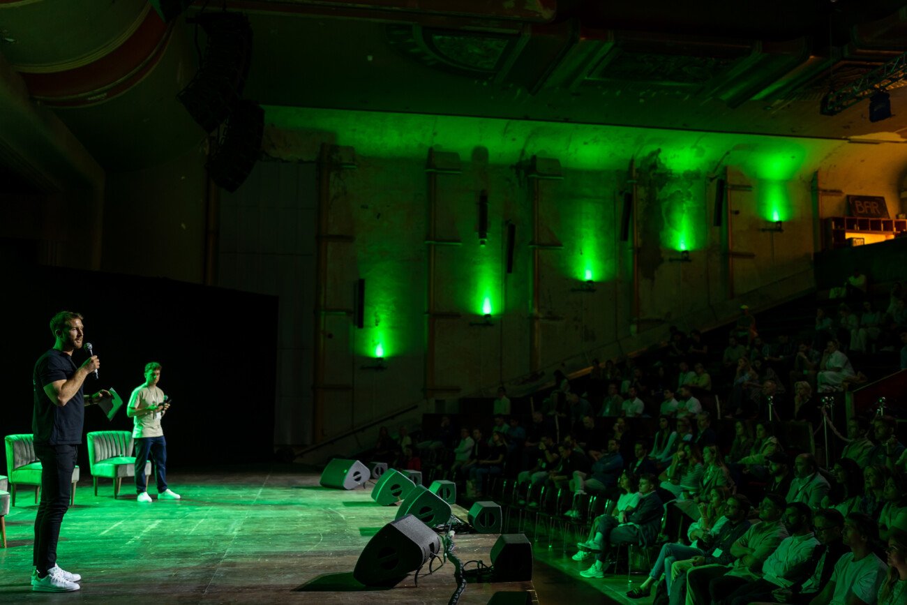 Two people stand on a dimly lit stage addressing an audience. The venue is decorated with green lighting on the walls. There are three empty chairs and some audio equipment on the stage. The audience is seated and attentive
