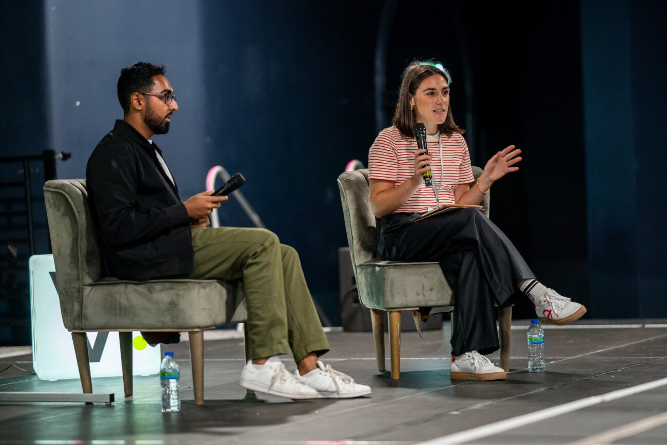 A man and a woman sit on stage in armchairs, holding microphones. The man wears a black jacket and green pants, while the woman wears a striped shirt and dark pants. Both have casual white sneakers. A water bottle is on the floor nearby