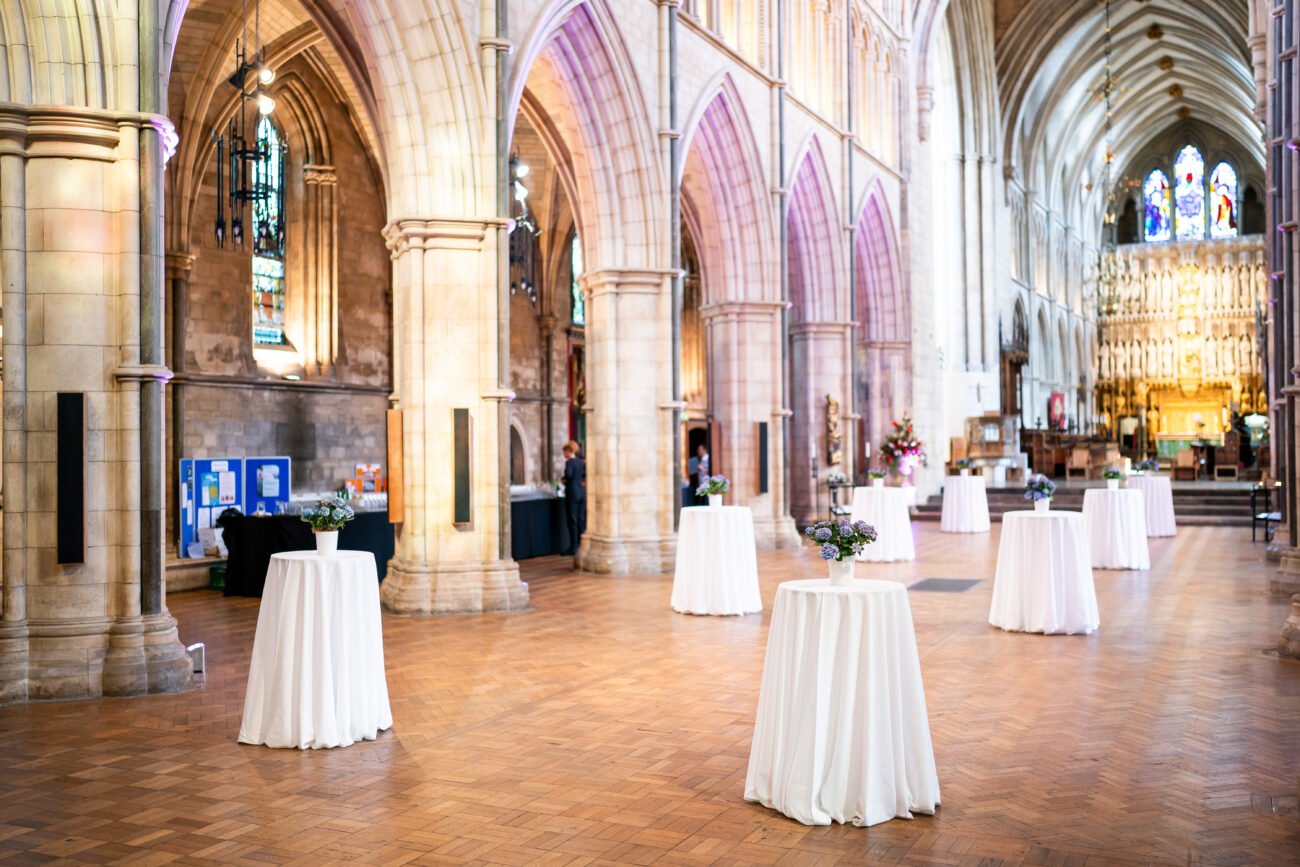 A spacious, ornate hall with tall arches and stained glass. Round tables with white tablecloths are placed throughout, each adorned with small floral centerpieces. Bright lighting highlights the architecture