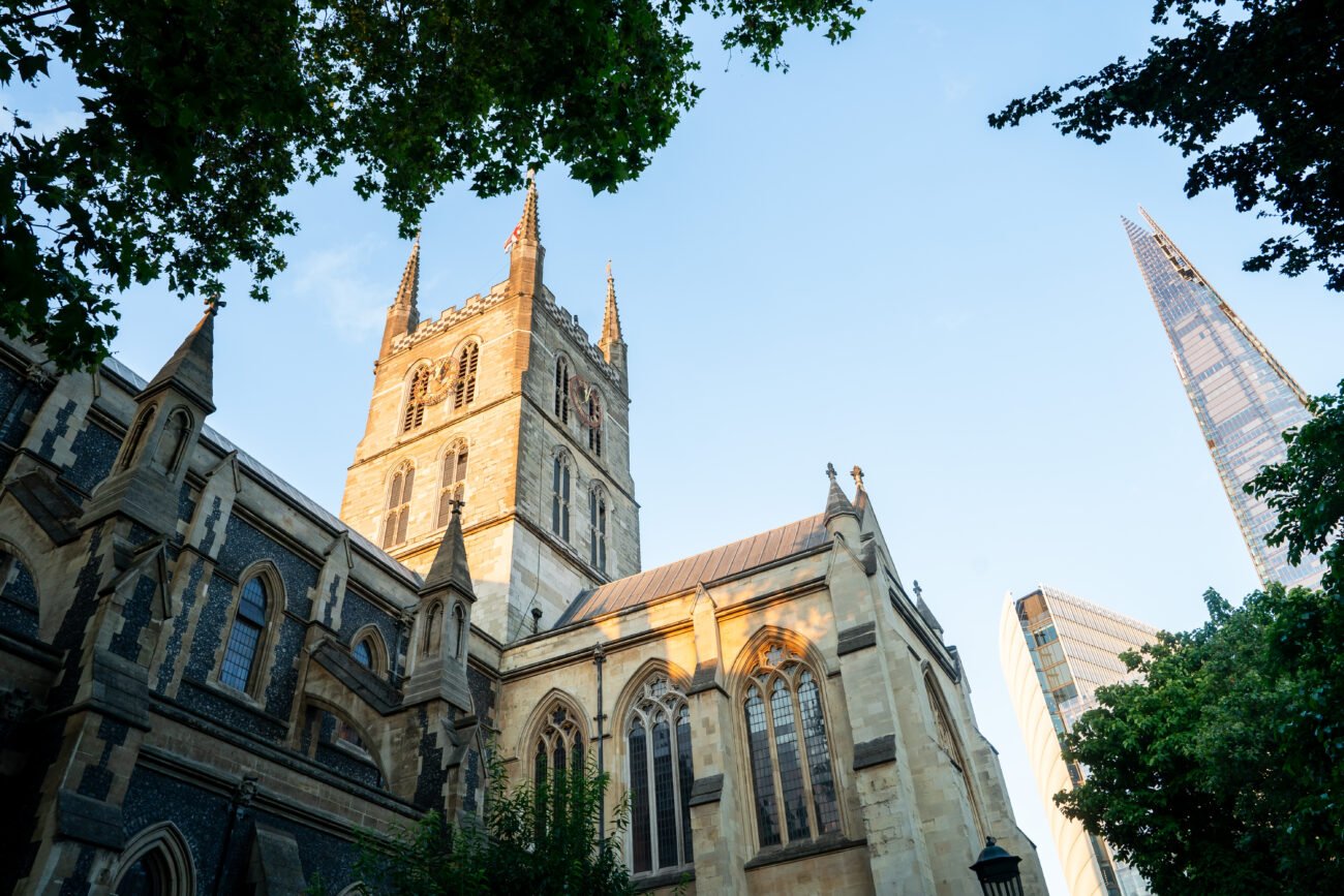 View of Southwark Cathedral with its Gothic architecture basking in sunlight, surrounded by trees. The modern Shard skyscraper is visible in the background against a clear blue sky