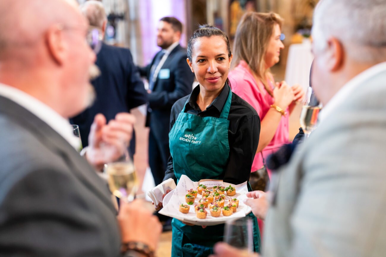 A smiling server wearing a green apron offers a tray of small appetizers to two guests at a social event. The guests hold wine glasses, and other attendees are visible in the background, engaging in conversation