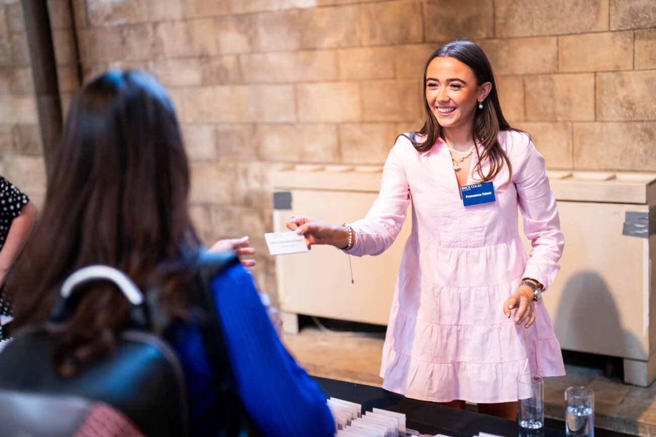 A woman in a pink dress and wearing a name tag is smiling and handing a card to another person across a table. The setting appears to be an indoor event with a stone wall in the background