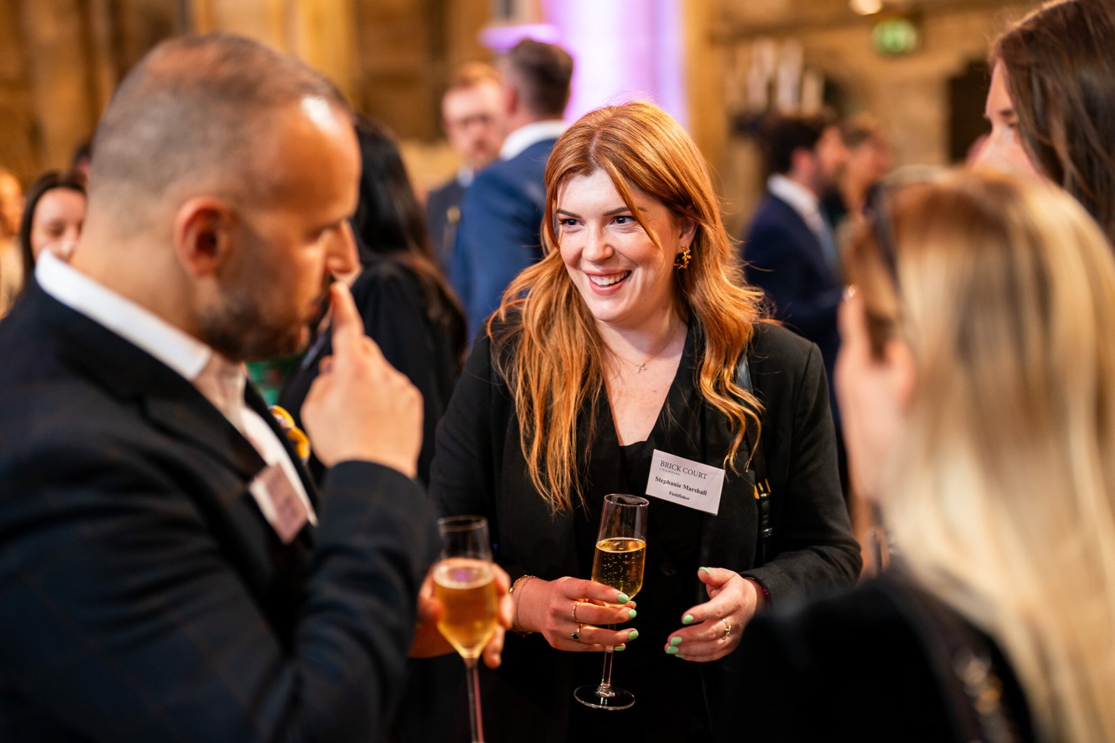 A group of people in formal attire are gathered at an indoor event, holding drinks and engaging in conversation. A smiling woman with long hair is in focus, wearing a name badge. The background is softly blurred, highlighting a lively atmosphere