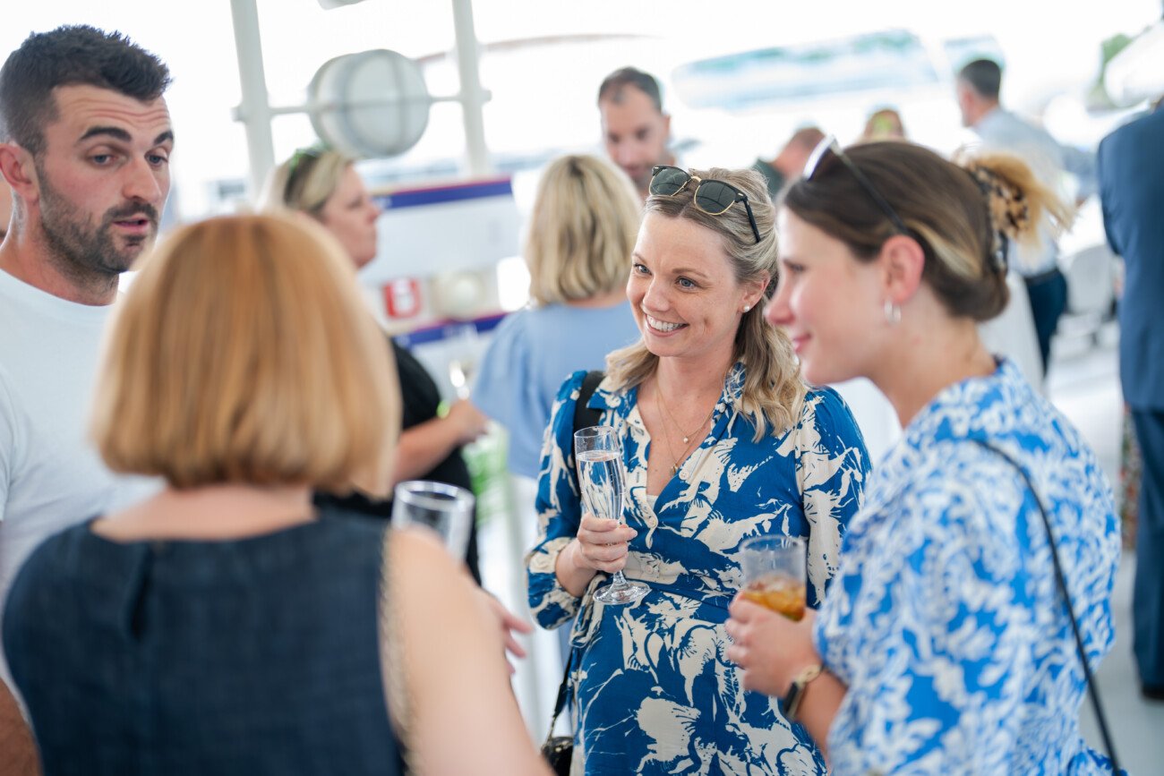 A group of people is socializing at a gathering at Lord Cricket Ground. They are holding drinks and engaged in conversation, effortlessly captured by event photography. The setting appears to be casual and indoor, with a relaxed atmosphere