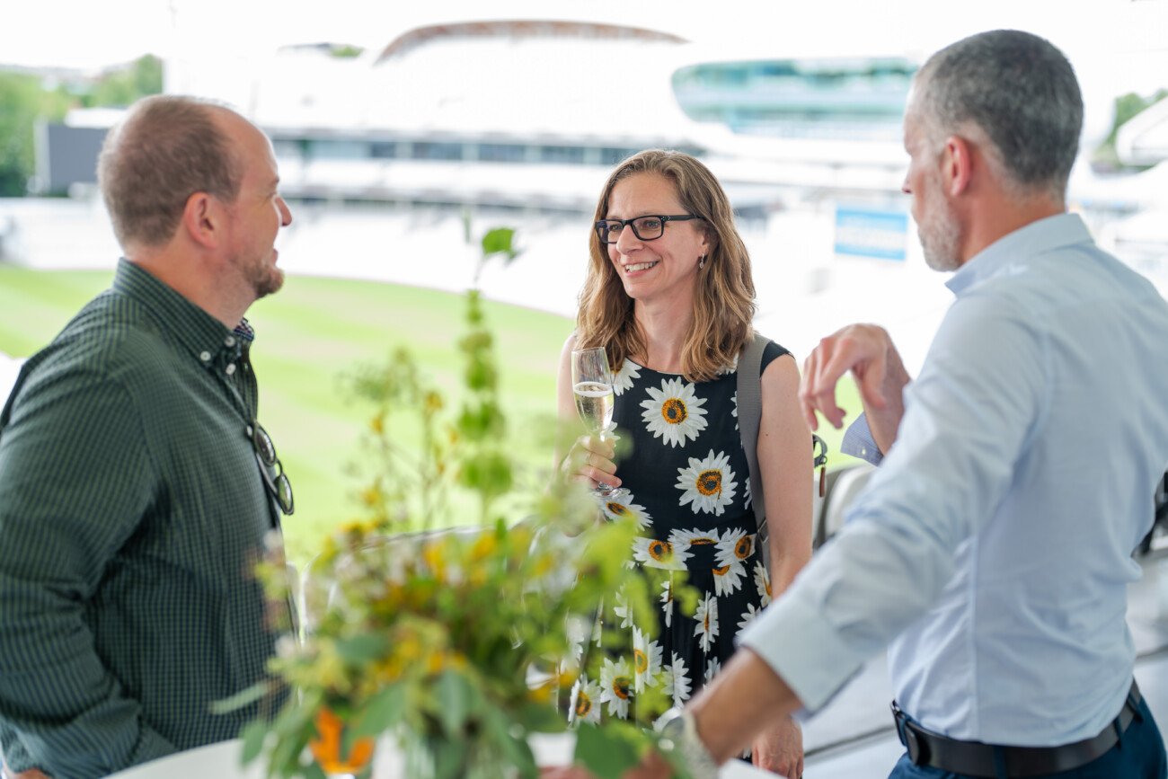 Three people stand outdoors at Lord Cricket Ground, engaged in conversation. The woman in the center, wearing a floral dress, holds a glass and smiles. The two men, one in a green checked shirt and the other in a blue shirt, appear to be listening. Blurry greenery is in the foreground—event photography perfection