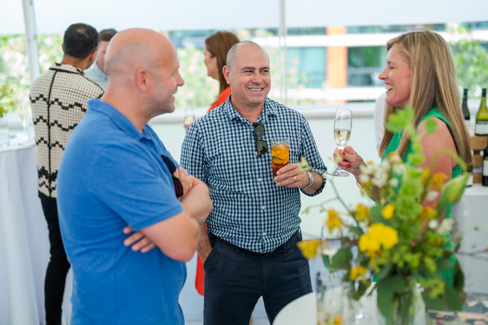Three people are engaged in conversation at an outdoor social event at Lord Cricket Ground. They stand near a table adorned with floral arrangements and drinks, one holding a glass with a beverage. The setting is perfect for event photography, capturing the casual and relaxed atmosphere