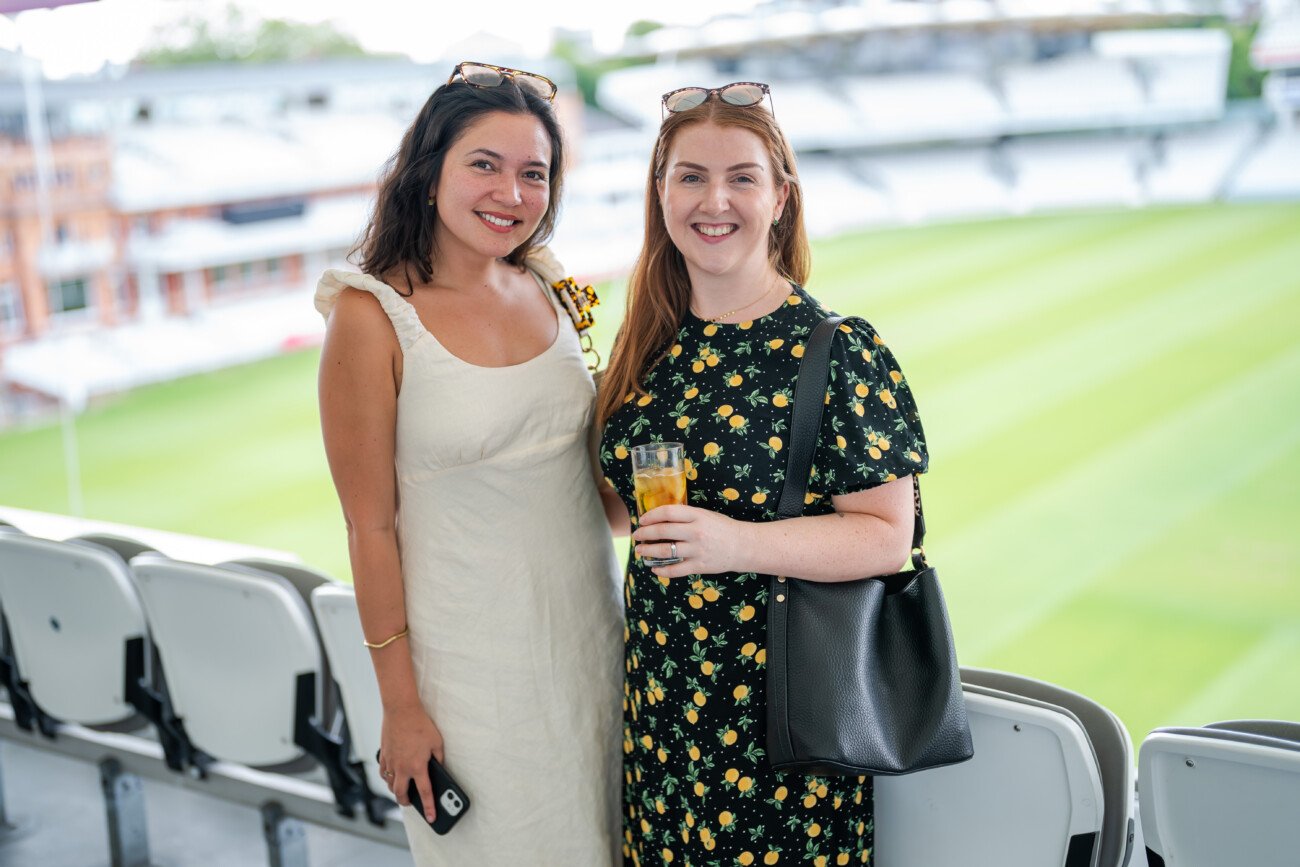 Two women, caught in the moment by event photography at Lord's Cricket Ground, smile in the stands. One wears a light dress with a phone in hand, while her friend, in a floral dress with a black bag and drink, poses against the backdrop of green grass and empty seats