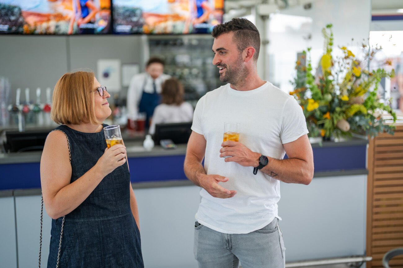 A woman and a man stand in a casual setting at Lord's Cricket Ground, drinks in hand, engrossed in conversation. The scene is the epitome of event photography, with a backdrop featuring people mingling and elegant floral arrangements, crafting a relaxed yet sophisticated atmosphere
