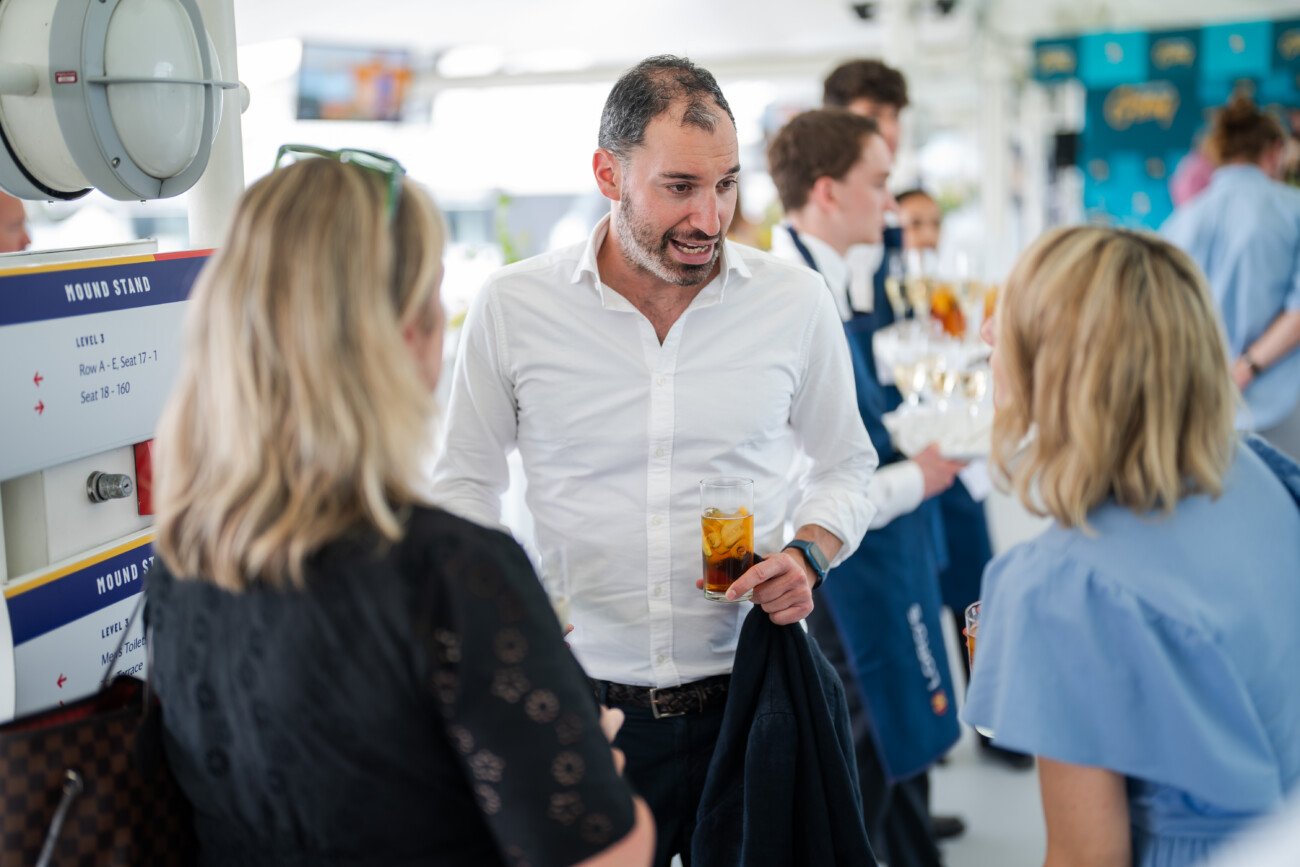 A man in a white shirt holds a drink and chats with two women at an indoor gathering, captured brilliantly in this example of event photography. In the background, attendees and servers bustle about, while a seating information sign hints that the setting is none other than Lord's Cricket Ground