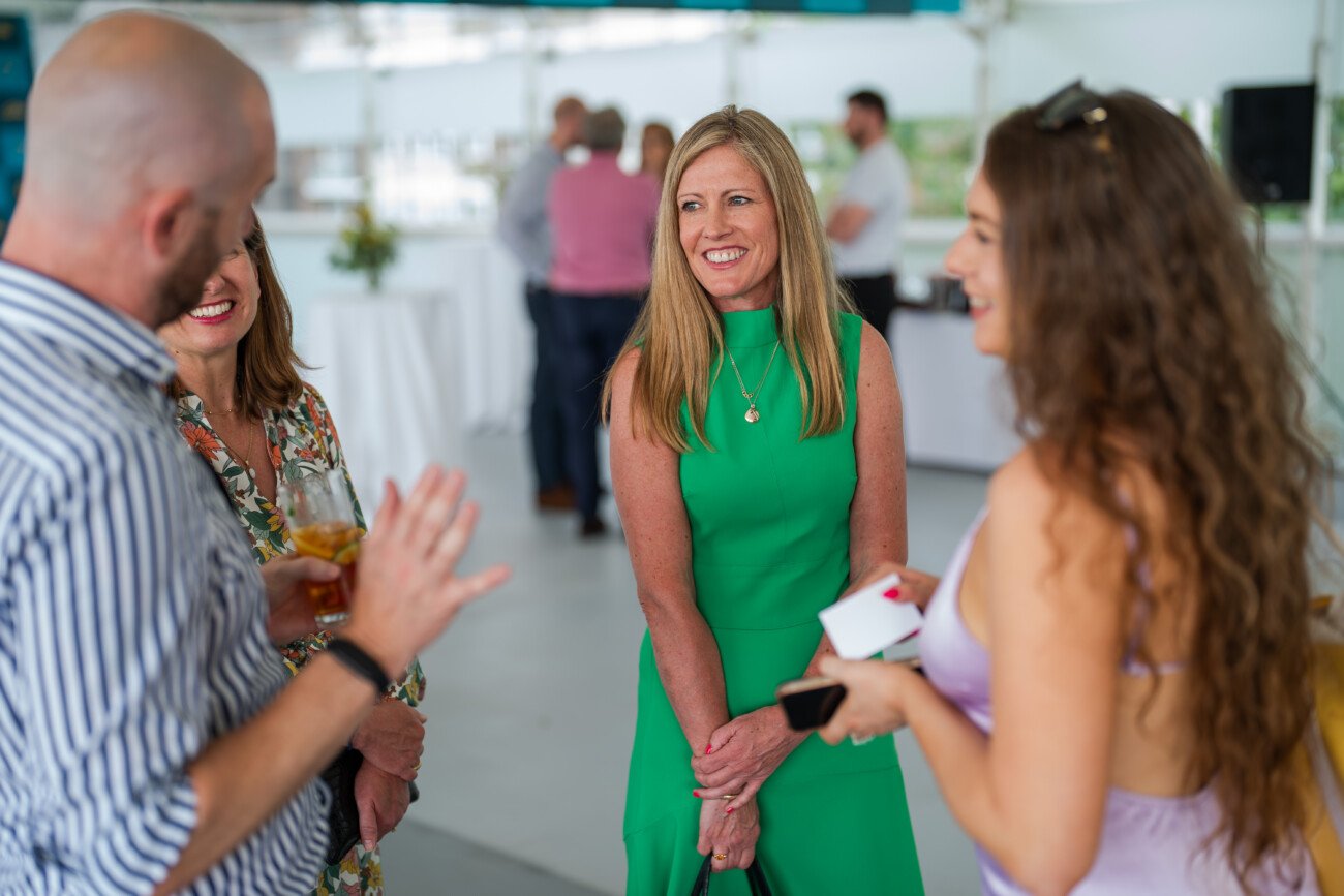 At a lively gathering at Lord's Cricket Ground, four people engage in spirited conversation. Two women and a man are captivated by a woman in a green dress, who smiles radiantly. In the background, event photography captures tables and the soft blur of other attendees enjoying the evening