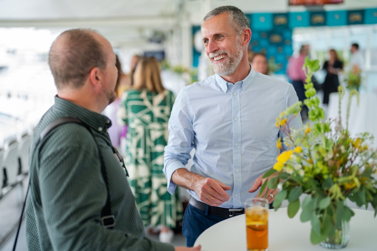 Amidst the vibrant ambiance of Lord Cricket Ground, two men share a laugh at a social event. One sports a light blue shirt, the other a dark green one. A floral arrangement and a glass with beverage grace the table between them, while blurred attendees lend depth to this moment of event photography
