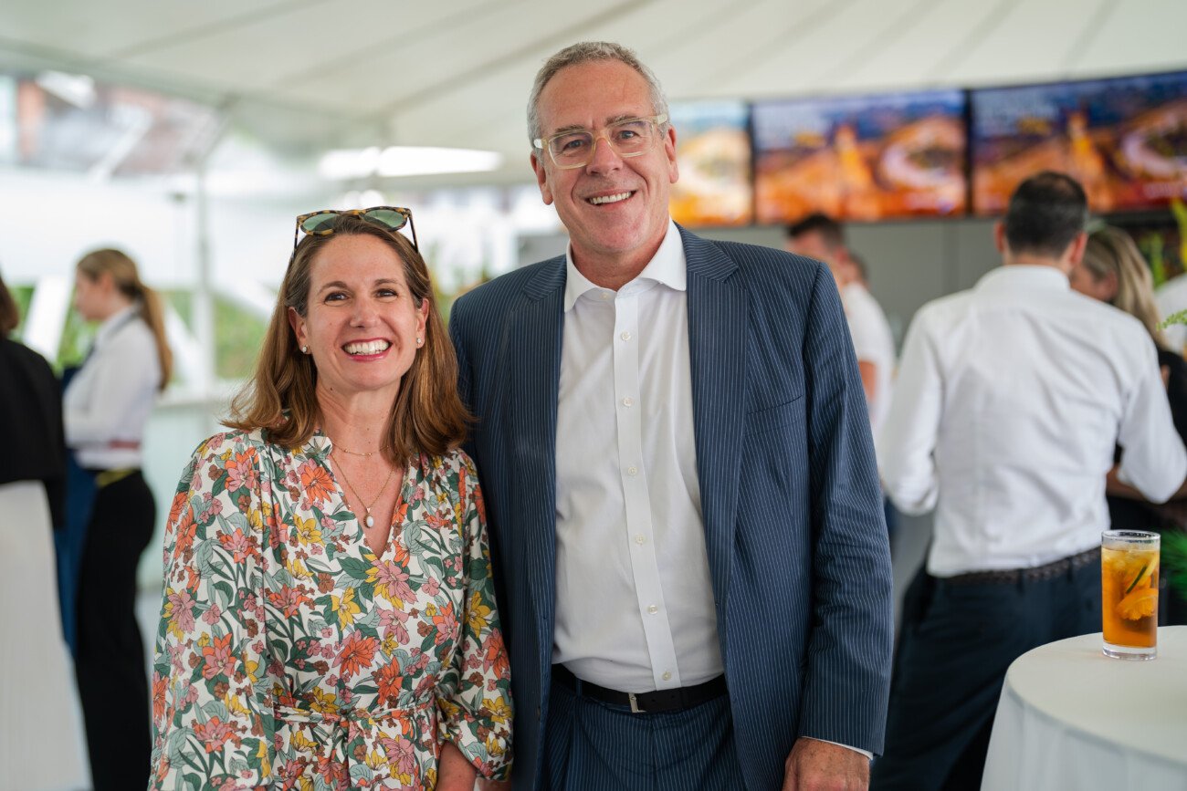 At an event at Lord's Cricket Ground, a woman in a floral dress and a man in a suit beam under the white tent. In the lively background, guests mingle amidst screens showcasing images, while a drink patiently sits on the round table nearby, capturing perfect moments with event photography magic