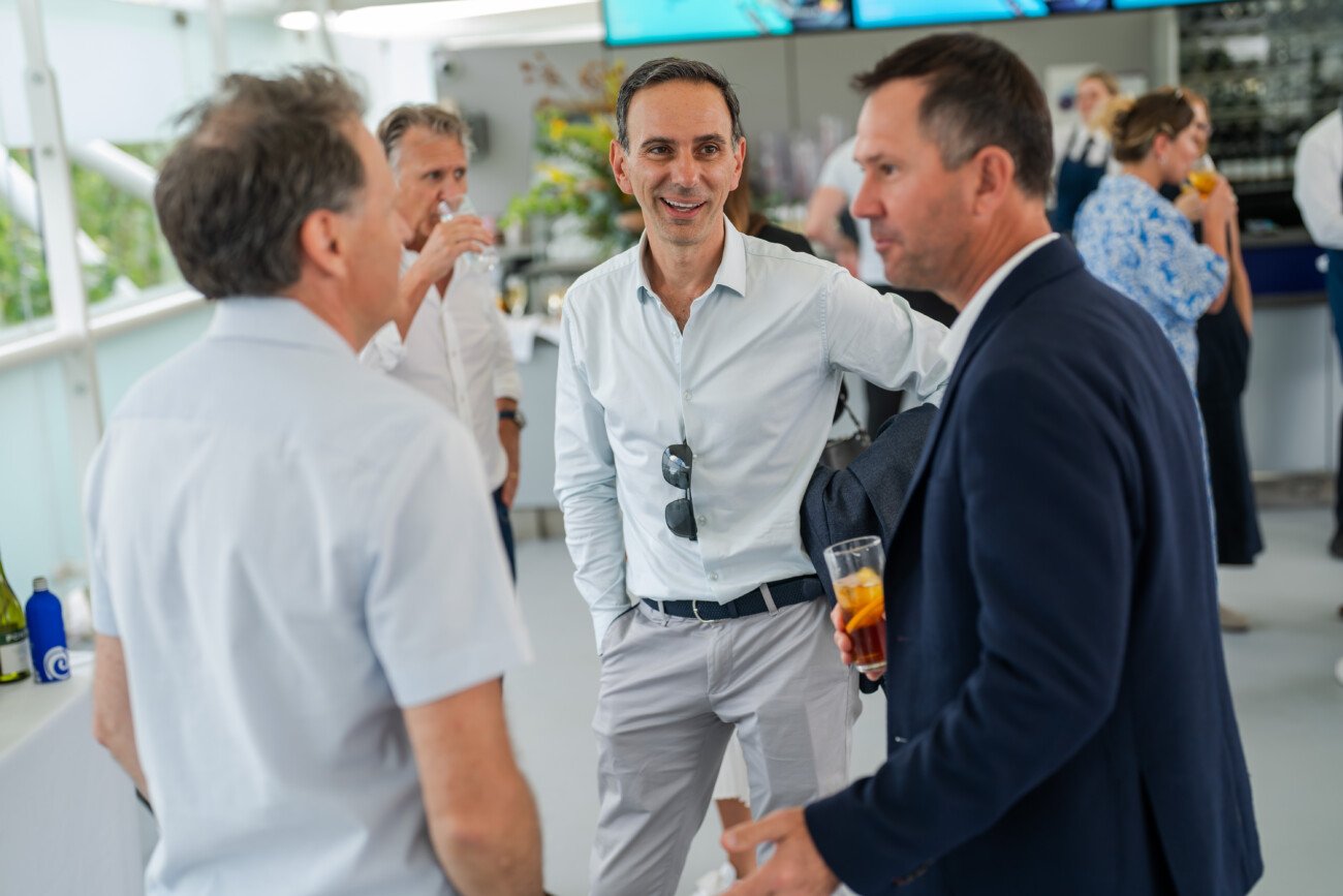 A group of people in semi-formal attire are captured with impeccable event photography at Lord's Cricket Ground. One person in a light shirt and sunglasses hangs from his collar while holding a drink, as others socialize near the bar in the background