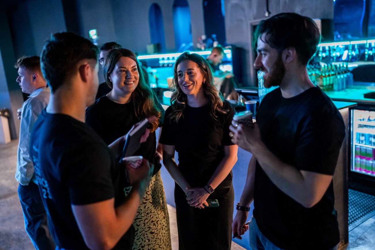 A group of four people stands in a bar, engaging in conversation. Two men hold drinks, while two women listen and smile. The background features neon lights and shelves filled with bottles, creating a lively atmosphere