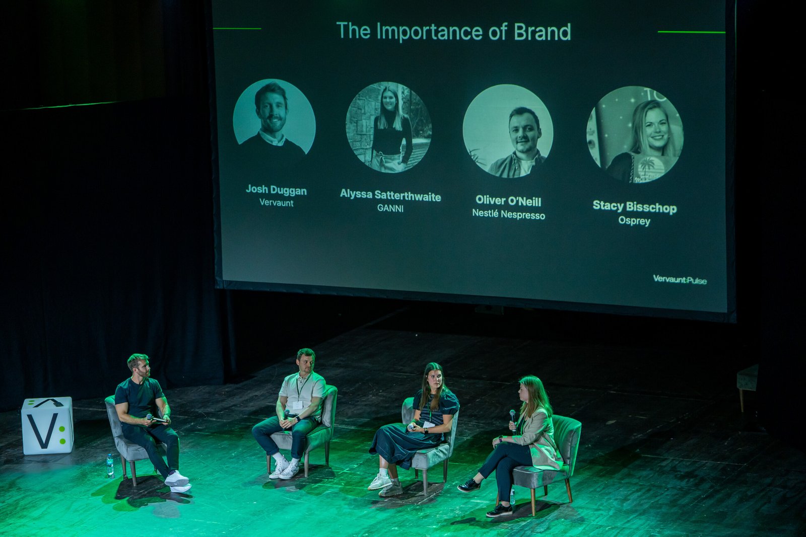 Four people are seated on a stage in front of a large screen labeled "The Importance of Brand," showing four headshots and names. The setting appears to be a panel discussion or presentation