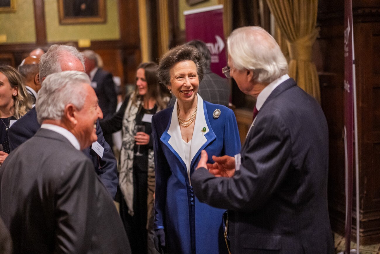 A woman in a blue coat smiles and converses with several people in a formal setting, captured by a London conference photographer. The room's wooden paneling and portraits set the scene, while other guests gather to add to the lively atmosphere
