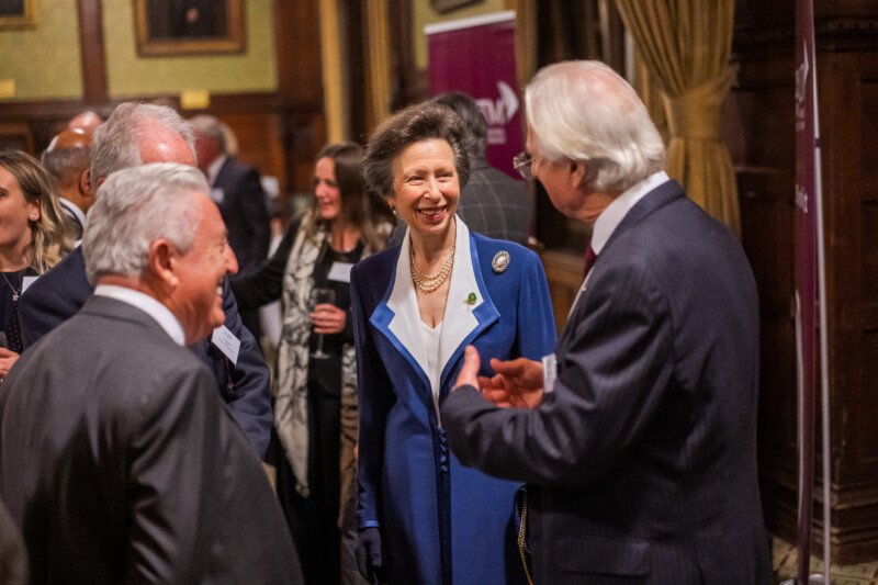 A woman in a blue coat smiles and converses with several people in a formal setting, captured by a London conference photographer. The room's wooden paneling and portraits set the scene, while other guests gather to add to the lively atmosphere