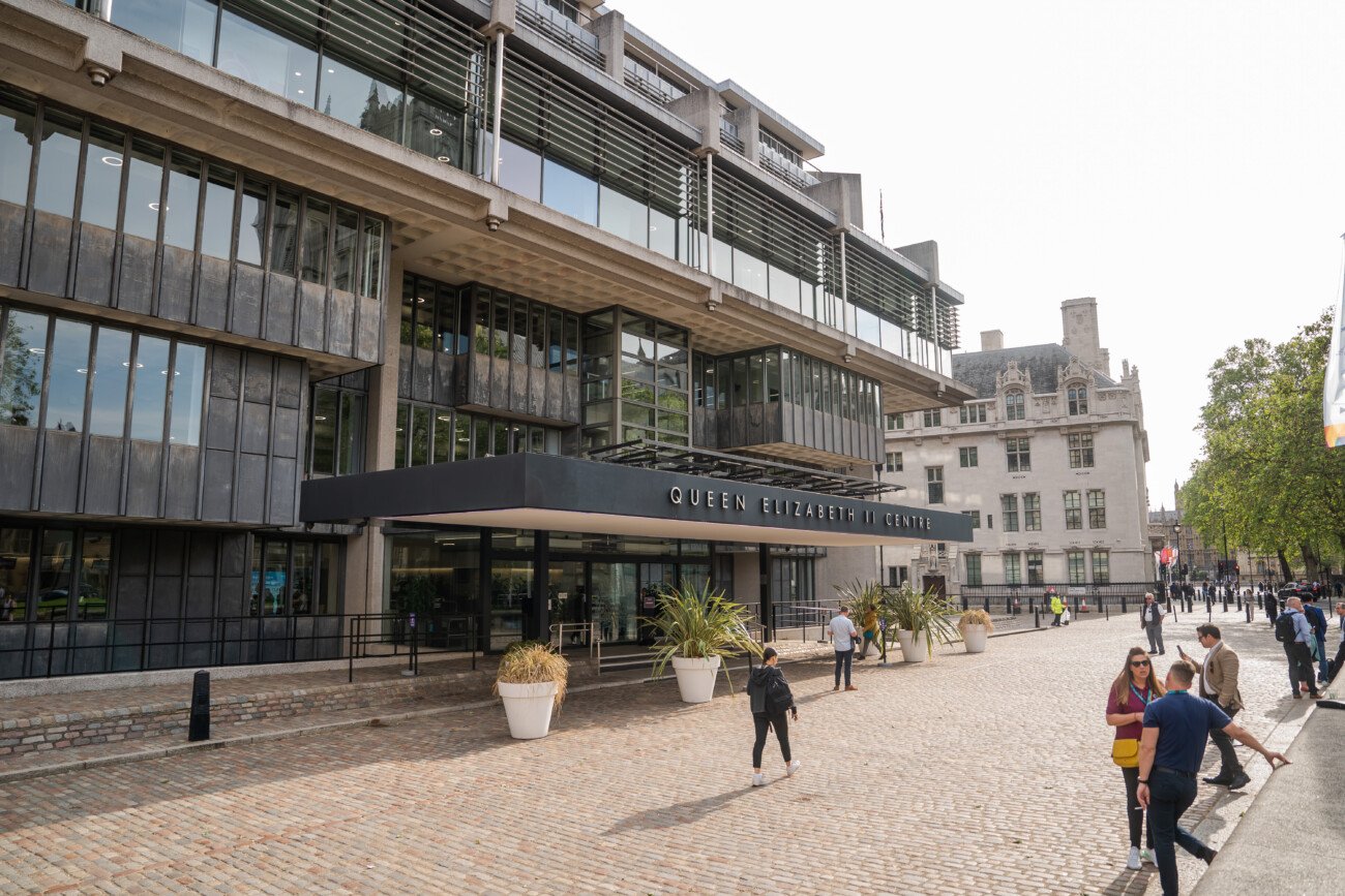 Street view of the Queen Elizabeth II Centre with a modern glass and concrete facade. People are walking on the cobbled path in front. The sun is shining, casting shadows, and greenery is visible in the background