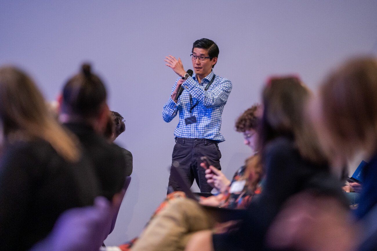 A man in a checkered shirt speaks into a microphone while standing among seated people, some of whom appear to be taking notes. The setting suggests a conference or seminar