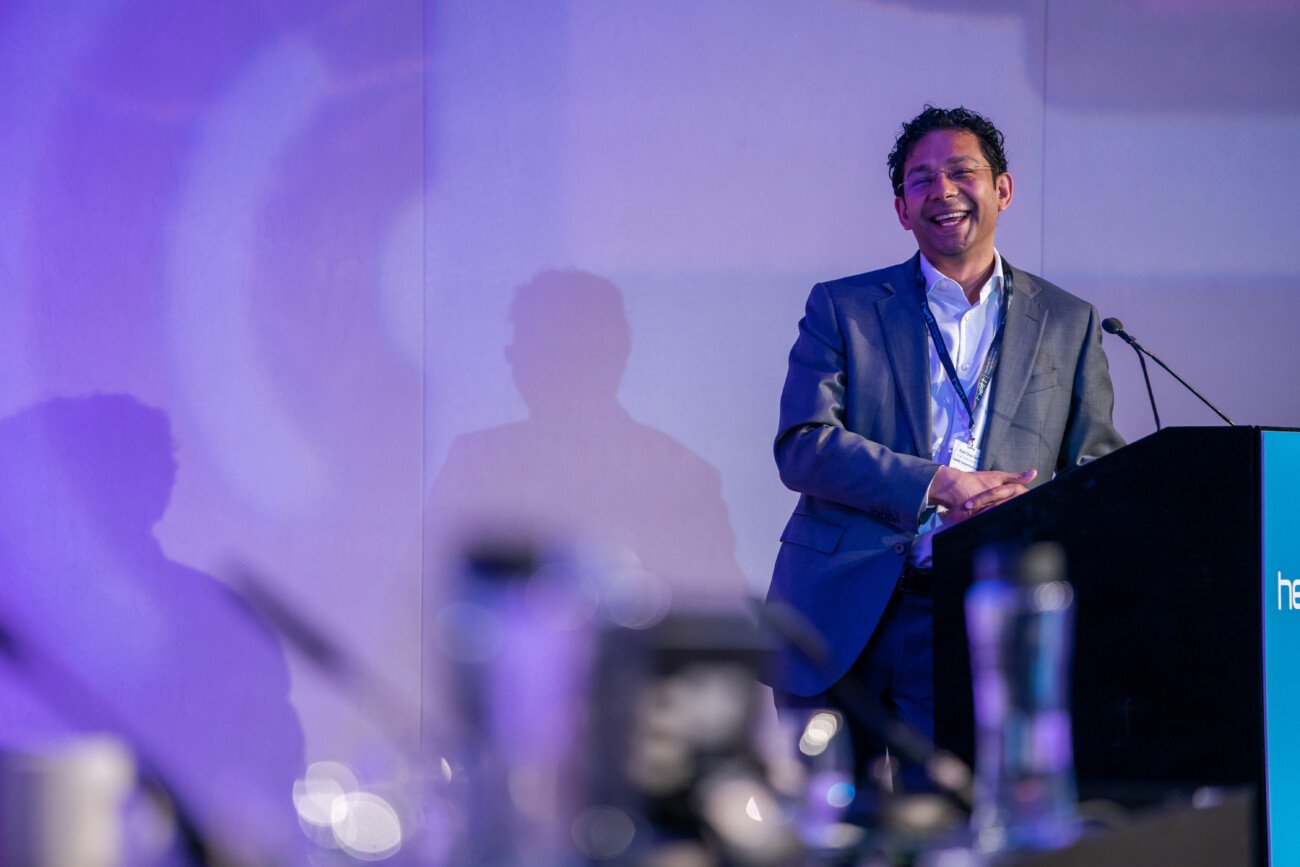 A man in a suit is smiling while standing at a podium during a presentation. The background has a purple hue, and there are blurred objects in the foreground