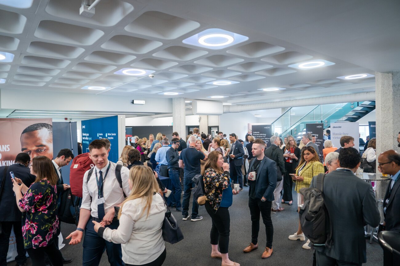 A large group of people engage in conversations at a networking event in a modern hall. Various promotional banners line the walls. Attendees are dressed in business casual attire, and some hold coffee cups. The ceiling features geometric lighting