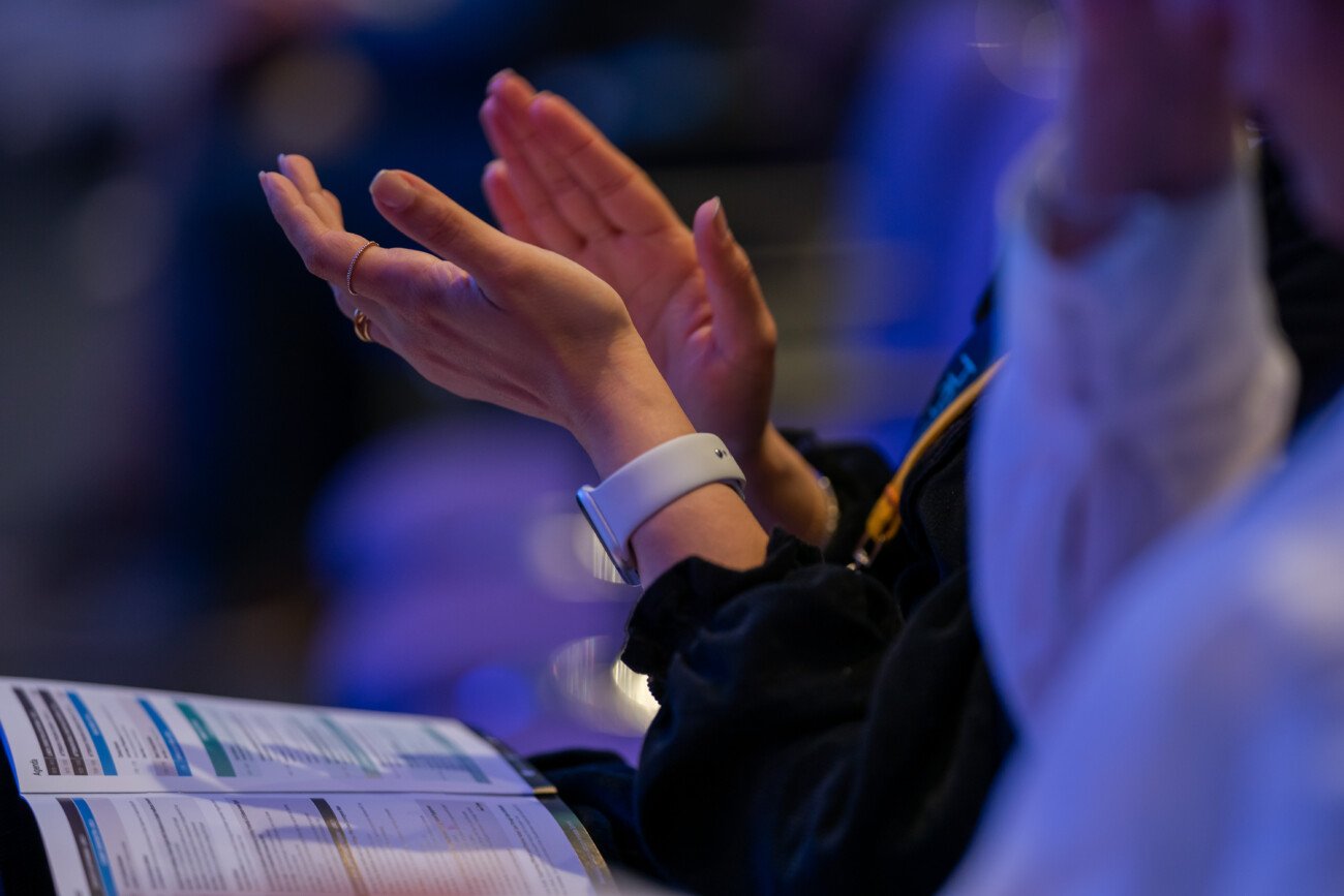 Person applauding while holding a program booklet on their lap. They are wearing a smartwatch and are seated in a dimly lit setting, suggesting a conference or event. The focus is on their hands and the paper, with a bluish ambient light