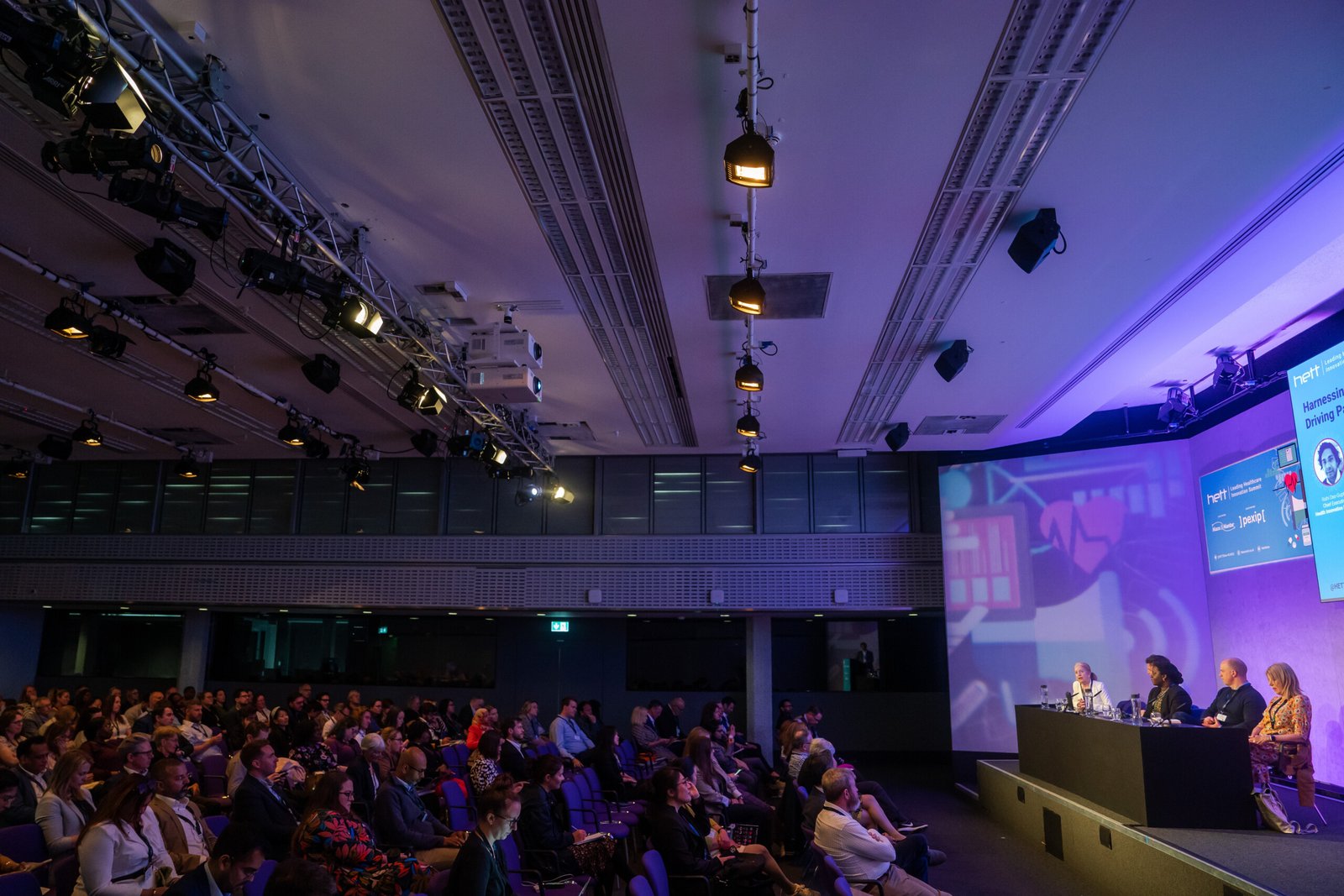 A panel of four people sits on a stage in a conference room with a large audience. The room is dimly lit, and a screen displays a presentation. The audience faces the stage, listening attentively