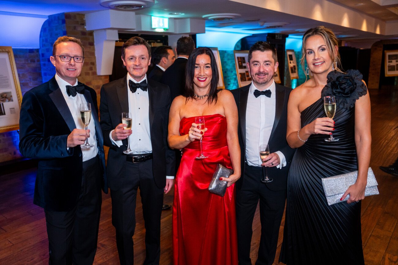 A group of five people, dressed in formal evening wear, are standing indoors holding glasses of champagne. The two women are wearing elegant gowns, and the three men are in tuxedos. The setting appears to be a formal event or gathering