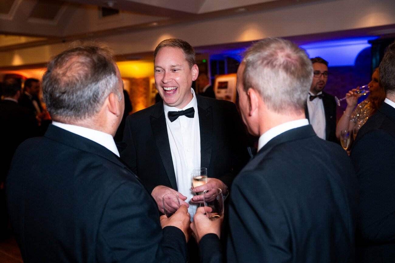 A group of people in formal attire, including a man smiling and holding a glass, are gathered at a social event. They appear to be engaged in conversation. The room is warmly lit, with colorful lights in the background
