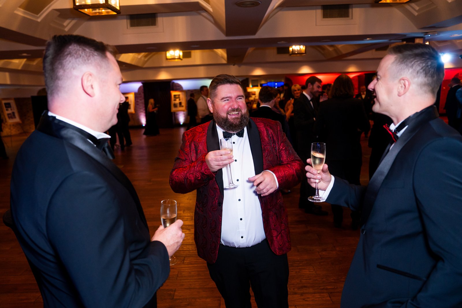 Three men in formal wear enjoy drinks and conversation at an elegant indoor event. One man wears a striking red patterned jacket, while the others wear black tuxedos. The warmly lit room is bustling with people in the background
