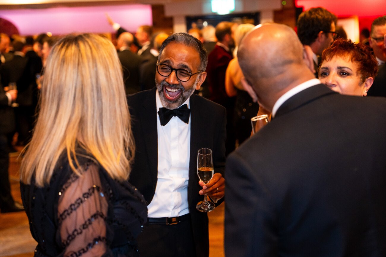 A group of people in formal attire are at a lively social event. A person in a tuxedo and bow tie is laughing while holding a champagne glass. Others around them are engaged in conversation, with blurred guests in the background