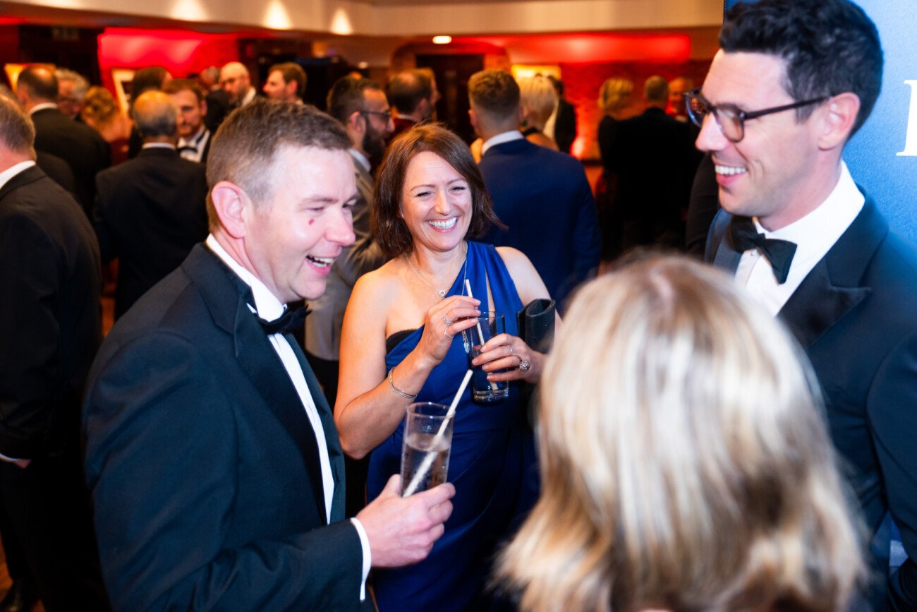 A group of people in formal attire enjoying a lively conversation at a social event. A man and a woman are holding drinks and smiling, while another man, wearing glasses, is laughing. The background is bustling with other guests