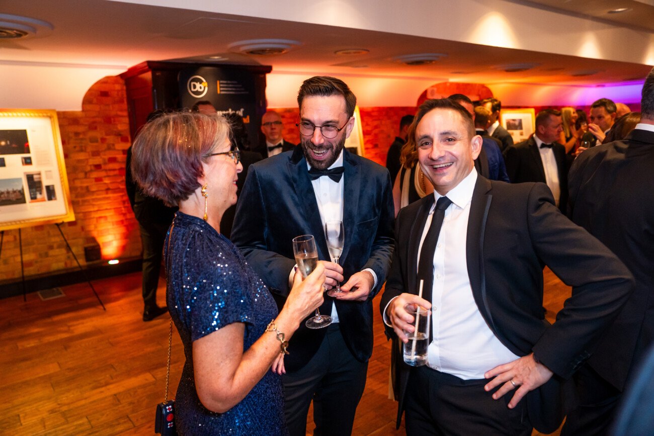 Three people in formal attire are smiling and conversing at an indoor event. One person is holding a champagne glass. The background features a framed picture, brick walls, and warm lighting