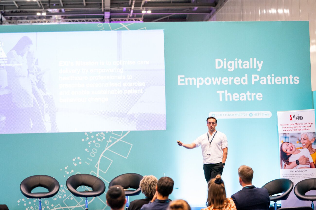 A speaker stands on a stage labeled "Digitally Empowered Patients Theatre," presenting to an audience. A large screen displays a presentation beside him. Several empty chairs are arranged in the foreground