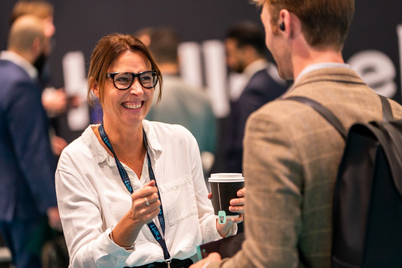 A woman in a white shirt and glasses smiles while holding a coffee cup and talking to a man in a brown suit. The background shows other people in business attire, suggesting a networking or professional event
