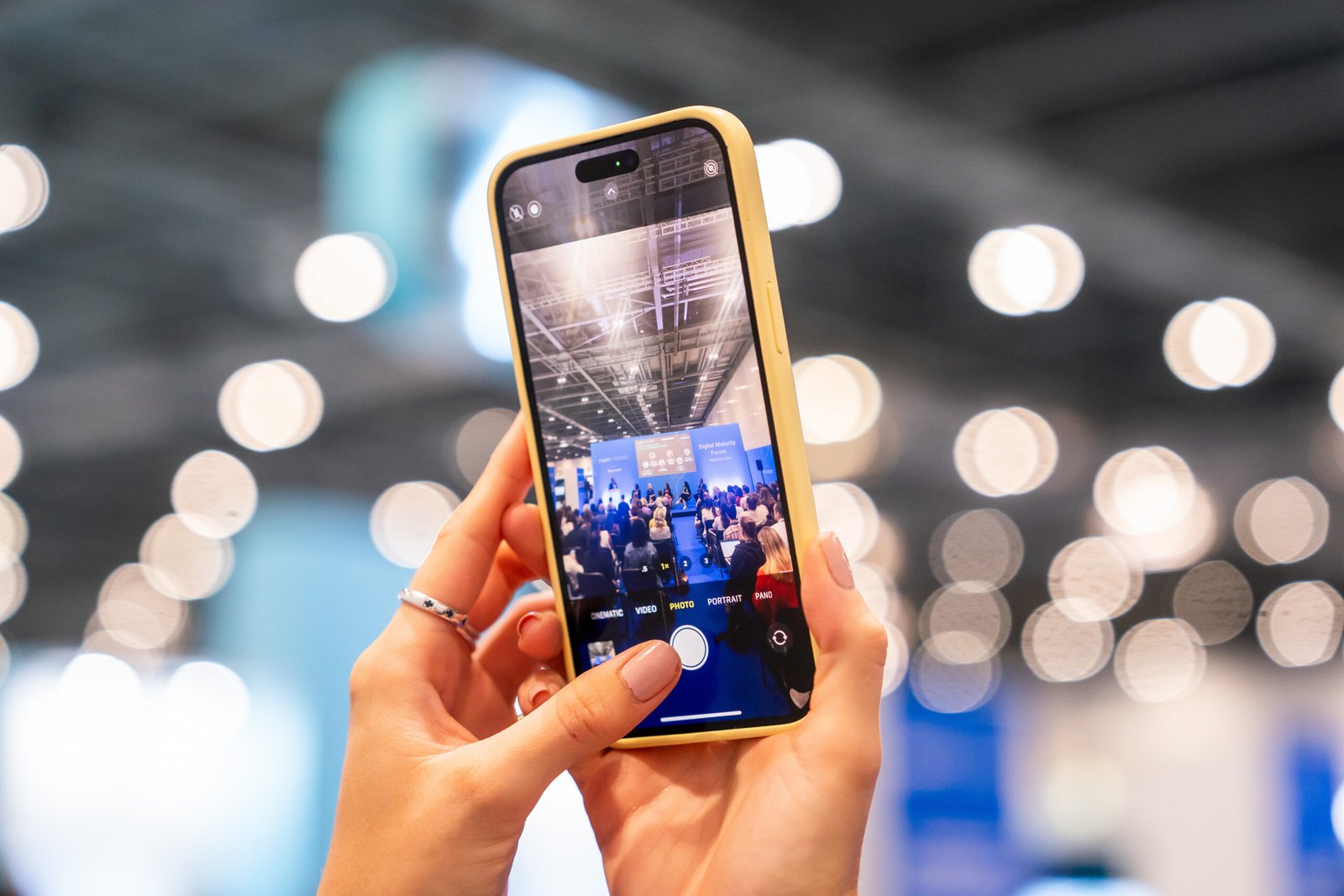 A person is holding a smartphone with a yellow case, capturing a video or photo of a conference or event. The background is blurred with bokeh lights and an audience visible on the phone screen
