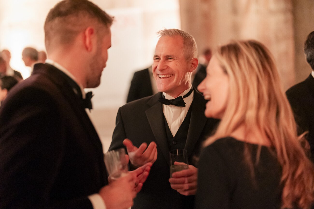 Three people in formal attire are engaged in conversation at a social event. The atmosphere is lively, with people in the background. The man in the center is smiling and holding a drink, as are the others
