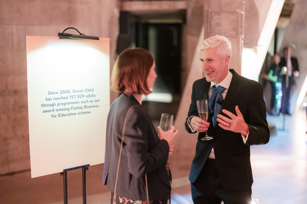 Two people in formal attire are talking and holding drinks. A sign beside them reads about Street Child's impact, mentioning 157,929 adults reached through its Family Business for Education scheme since 2008