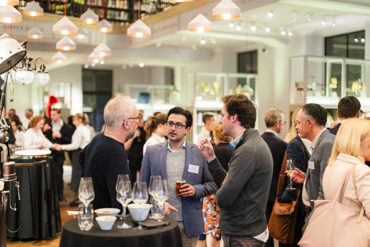 People are gathered at a networking event in a well-lit, modern space. Groups of attendees are engaged in conversation, holding drinks. The room is decorated with hanging lights, and there are tables with glasses and bowls