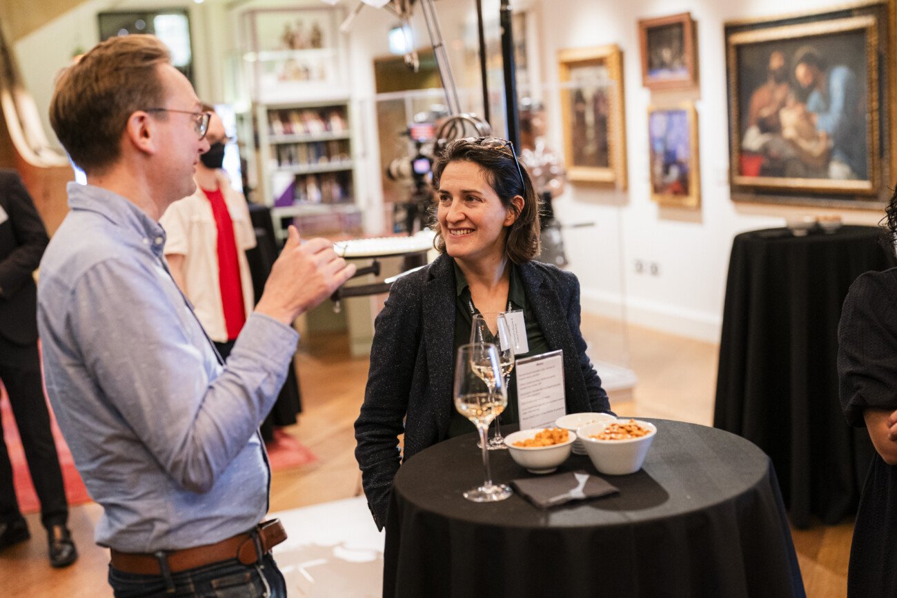 Two people engaged in conversation at a social event. A woman sits at a round table with snacks and a glass of wine. The man stands beside her gesturing while talking. Artwork and books are visible in the background