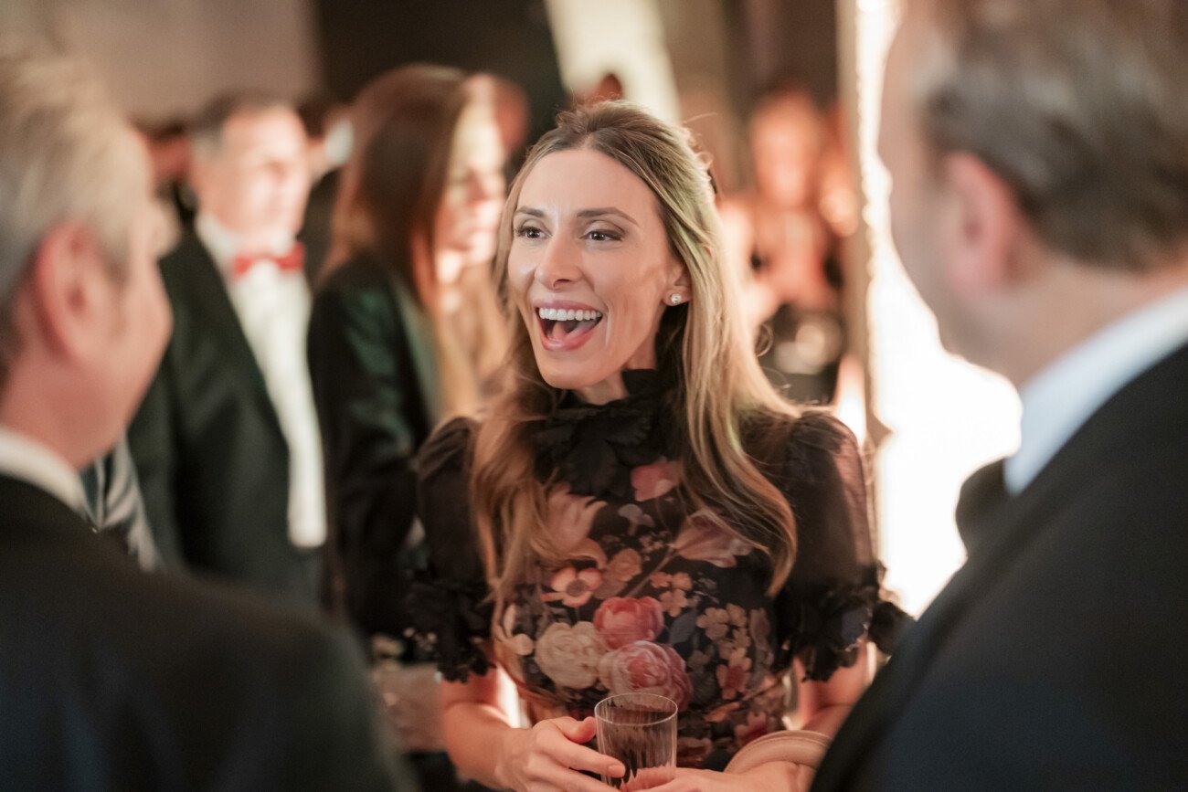A woman in a floral dress smiles and holds a drink while talking with two men in suits at a social gathering. The background is softly lit, and other people are visible but out of focus