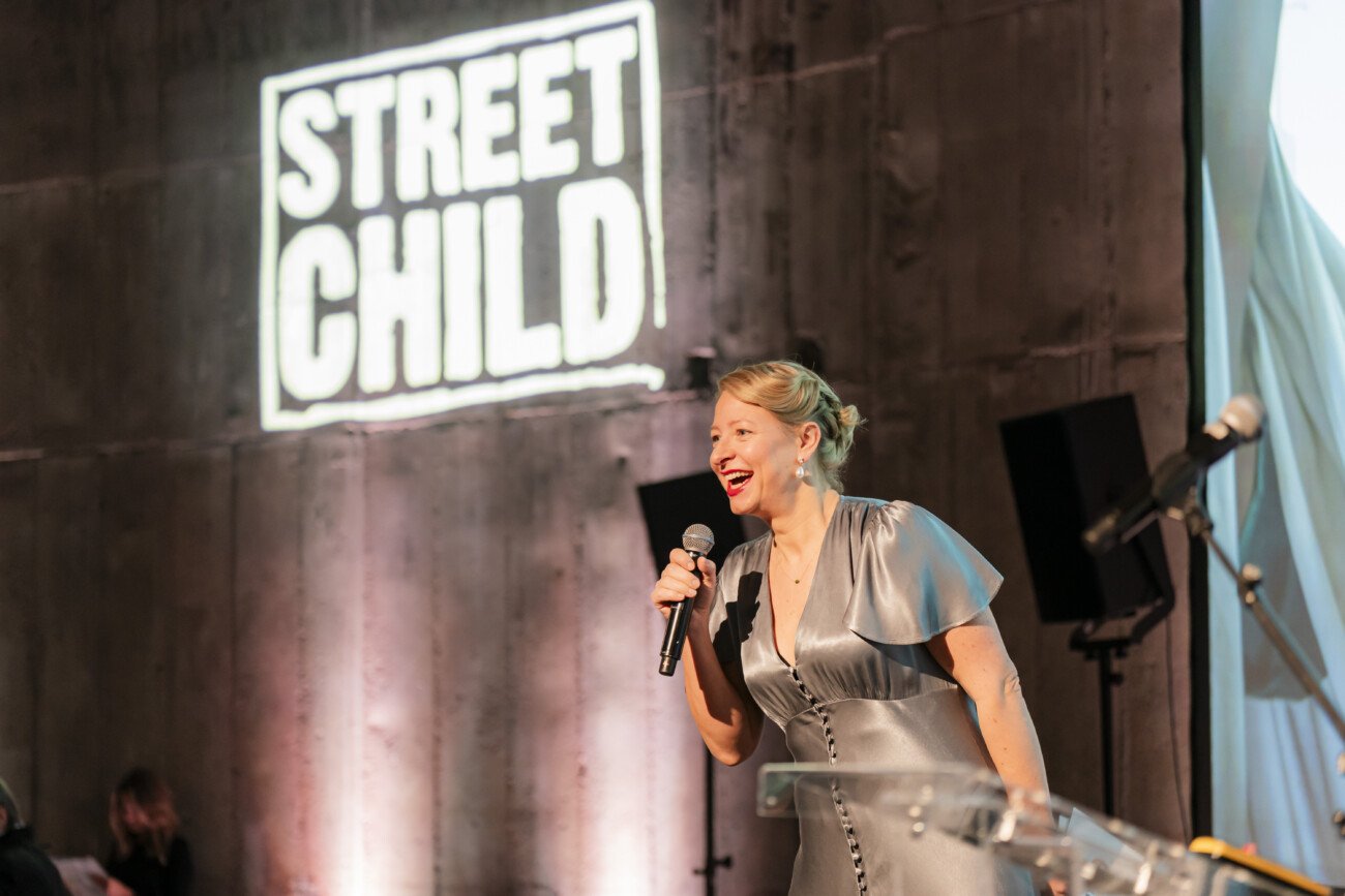A person in a silver dress speaks into a microphone on stage at an event. The backdrop features the "Street Child" logo on a concrete wall. Stage lights are visible