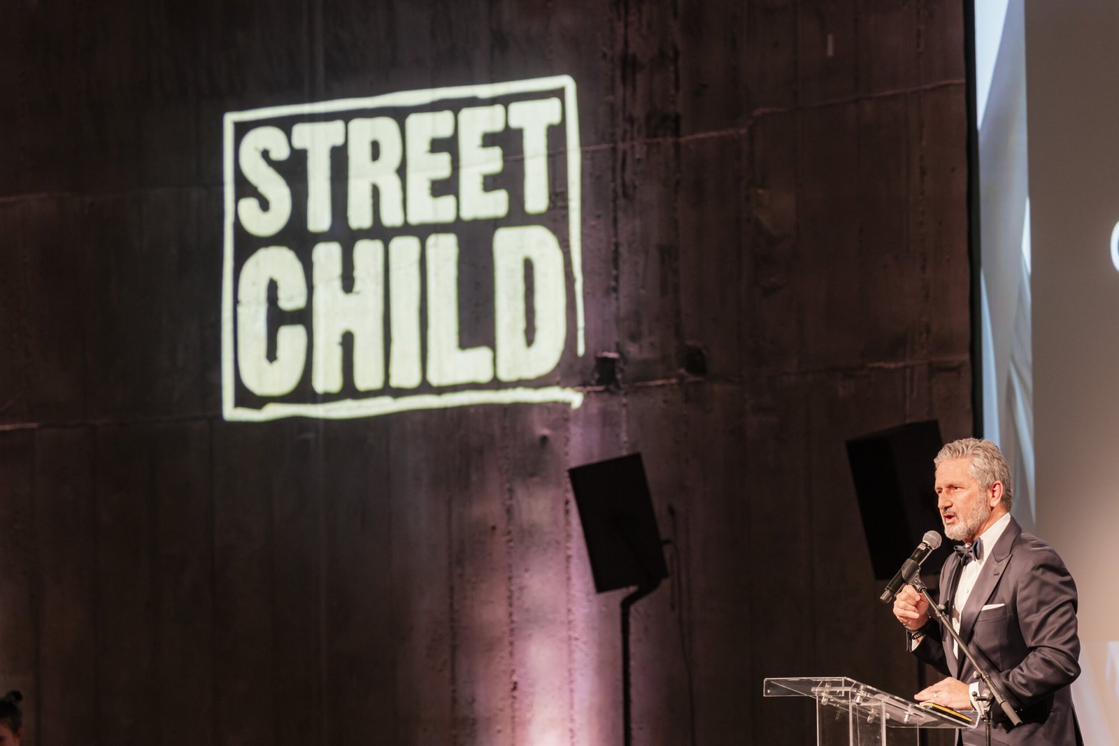 A man in a suit speaks at a podium in front of a textured wall with the illuminated logo "Street Child." The setting appears to be a formal event or conference