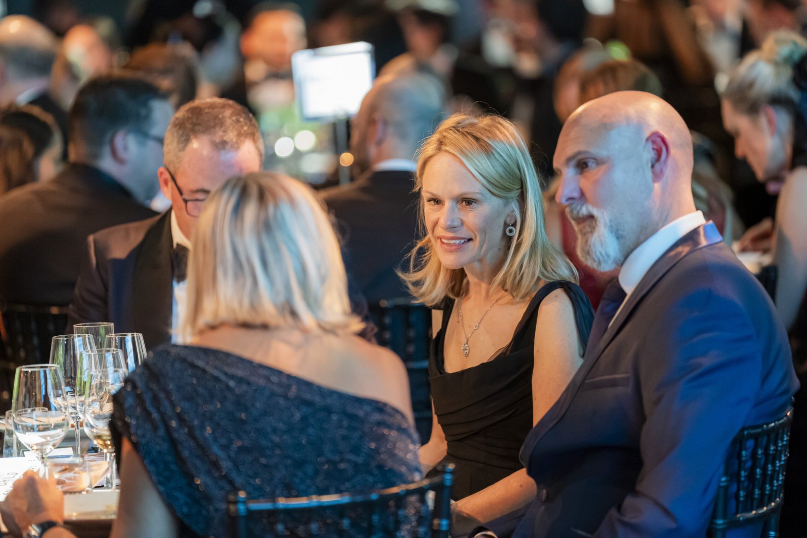 People in formal attire seated around a table at a gala dinner, engaged in conversation. The setting is elegantly lit, with glasses and plates on the table. The background shows other attendees blurred in soft focus. James Gifford-Mead Photography - Event Photographer London