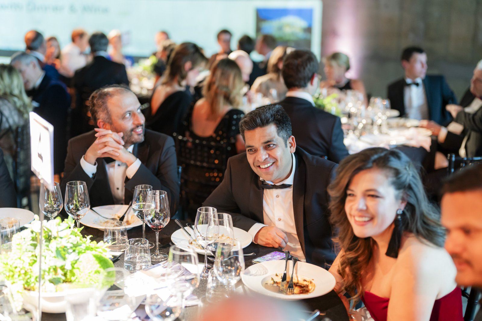 People dressed in formal attire sit at a table during an elegant dinner event. The table is set with wine glasses and plates. The atmosphere is lively, with guests engaged in conversation and smiling. James Gifford-Mead Photography - Event Photographer London