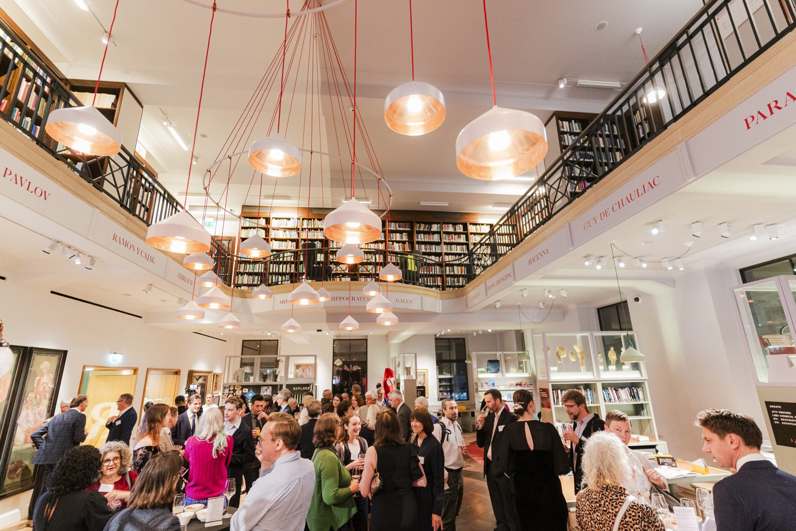A large group of people gathers in a spacious library with two levels of bookshelves. Warm, modern pendant lights hang from the ceiling, and framed art is displayed on the walls. The atmosphere is lively and social