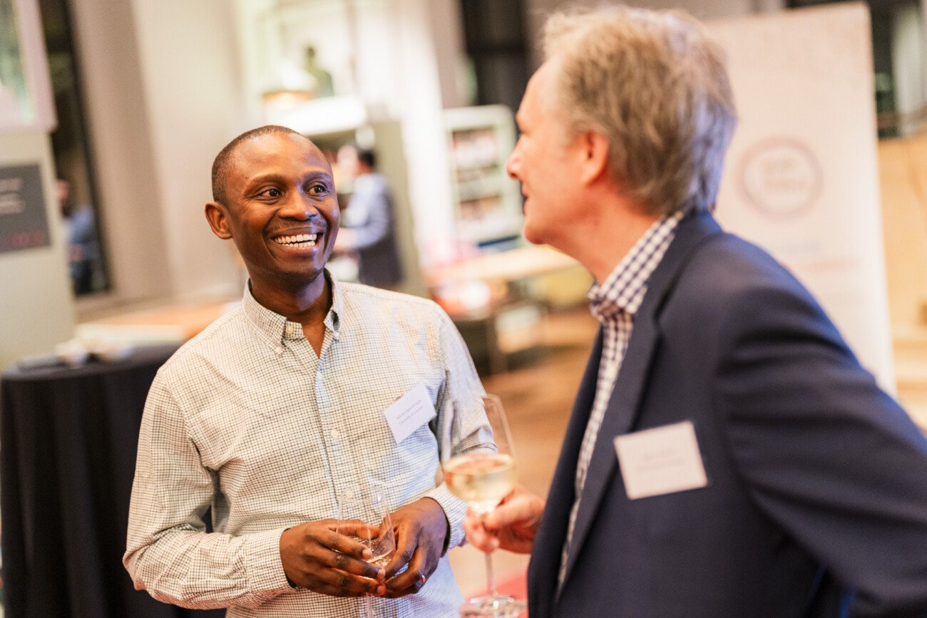 Two men in business attire are conversing at an indoor event. One, holding a beverage, is smiling, while the other listens attentively with a wine glass in hand. They are in a well-lit room with blurred background details
