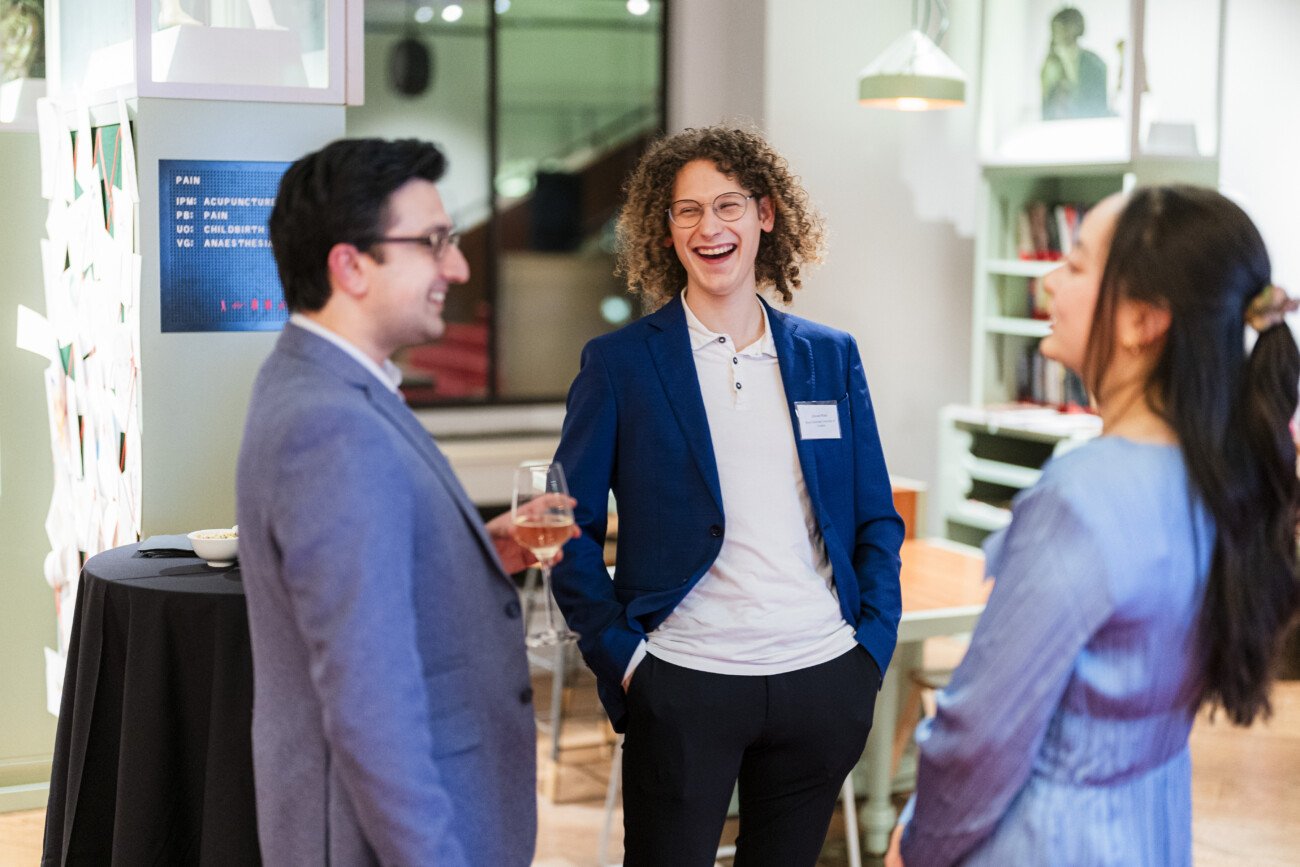 Three people in business attire stand and converse at a social event. One man, wearing a blue jacket, smiles broadly. They are indoors, with a table, chairs, and wall decor in the background